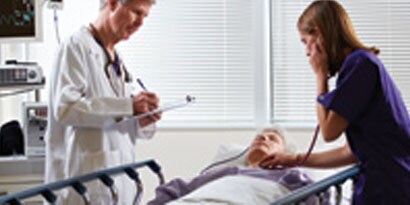 A doctor at a patients bedside, a nurse uses stethoscope on patient
