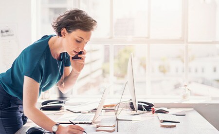 Woman on phone standing over desk taking notes in bright white sun-bathed office with large windows
