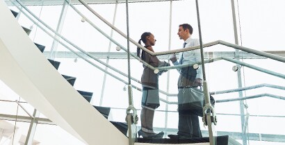 A man and a woman stop to talk on a stairway that is surrounded by floor-to-ceiling windows.
