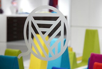 Clear glass window with etched glass-style line art logo with colorful shapes behind.
