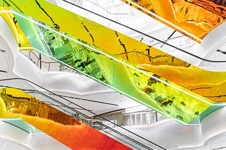 Multi-level mall with white walls and suspended escalators covered in colorful, reflective 3M Dichroic Glass Finishes with red, yellow, orange, and green tones.

