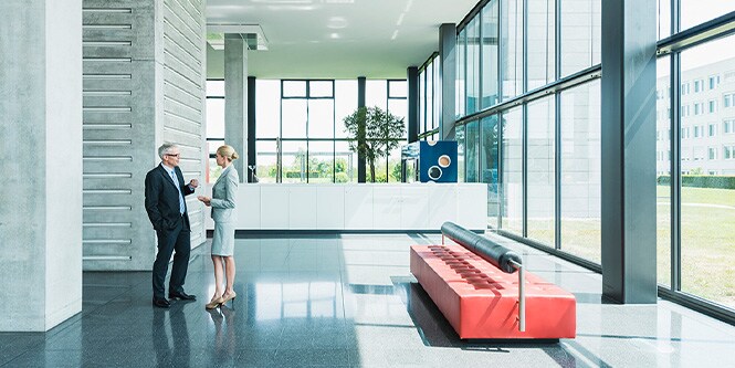 Wide shot of modern office building lobby surrounded by walls of windows with two businesspeople conversing

