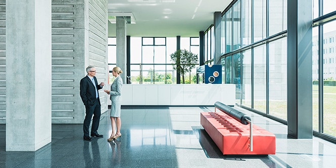 Wide shot of modern office building lobby surrounded by walls of windows with two businesspeople conversing
